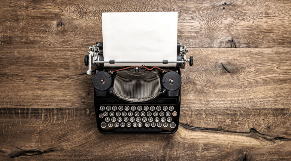 Eine klassische Schreibmaschine, eines der Retro-Gadgets, steht auf einer Holzoberfläche. Das Gerät hat schwarze Tasten mit weißen Buchstaben und ein eingespanntes leeres Blatt Papier, das den Charme des analogen Schreibens vergangener Zeiten widerspiegelt.