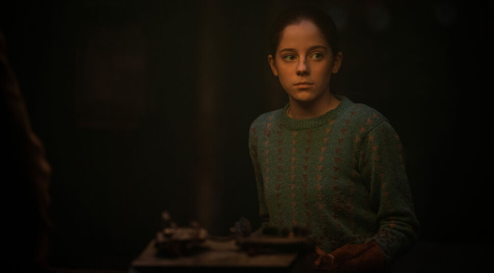 Auf dem Bild im Silo Staffel 3 Artikel ist die junge Juliette in einem Rückblick zu sehen. Sie trägt einen grünen, abgenutzten Pullover mit rotem Muster und schaut nachdenklich zur Seite. 