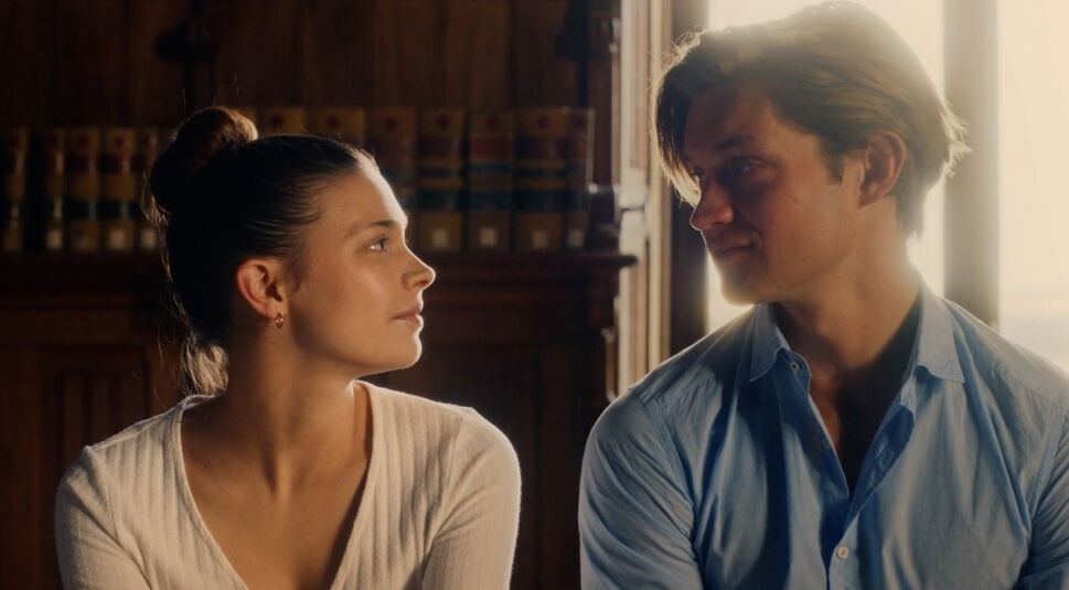 Auf dem Bild zu den besten Serien 2024 sind Ruby Bell, gespielt von Harriet Herbig-Matten, und James Beaufort, dargestellt von Damian Hardung, zu sehen. Die beiden sitzen nebeneinander, schauen sich tief in die Augen und wirken vertraut. Ruby trägt ein schlichtes weißes Oberteil, James ein hellblaues Hemd. Die warme Lichtstimmung und der Hintergrund mit alten Büchern schaffen eine romantische und ruhige Atmosphäre.