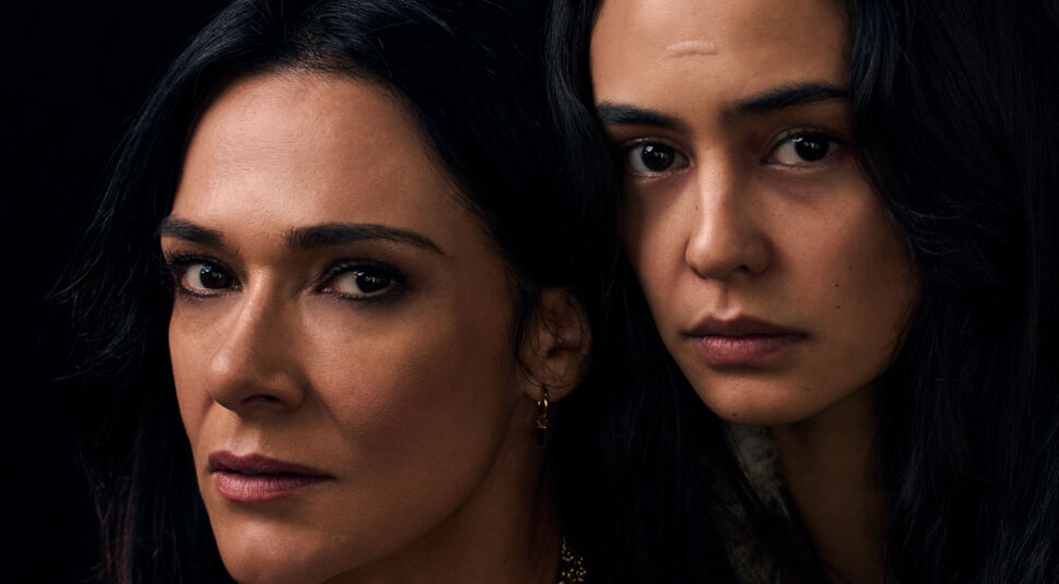Auf dem Bild zu Yellowjackets Staffel 3 sind Courtney Eaton und Simone Kessell als Lottie zu sehen. Ihre Gesichter sind nah beieinander, beide blicken mit ernsten, intensiven Augen in die Kamera. Simone Kessell trägt dezentes Make-up und goldene Ohrringe, während Courtney Eaton einen natürlichen Look hat. Die dunkle Kulisse verstärkt die geheimnisvolle und bedrohliche Atmosphäre der Szene.