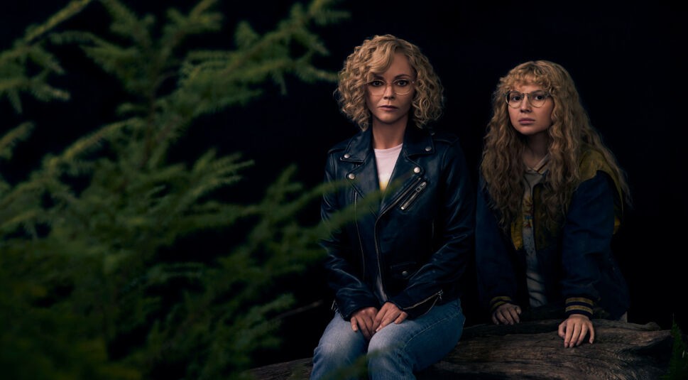 Auf dem Bild zu Yellowjackets Staffel 3 sind Christina Ricci und Samantha Hanratty als Misty zu sehen. Christina Ricci trägt eine schwarze Lederjacke und blickt ernst in die Kamera, während Samantha Hanratty in einer abgenutzten Jacke mit wilder Lockenmähne daneben sitzt. Beide tragen eine Brille, was ihre Verbindung als jüngere und ältere Version der Figur Misty unterstreicht. Die düstere Waldkulisse verstärkt die geheimnisvolle Atmosphäre.