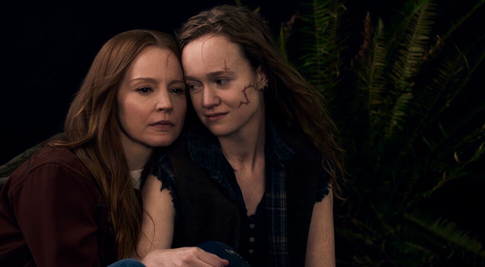 Auf dem Bild zu Yellowjackets Staffel 3 sind Lauren Ambrose und Liv Hewson als Van zu sehen. Sie sitzen eng beieinander in einer düsteren Waldumgebung. Liv Hewsons Van hat markante Narben im Gesicht und blickt sanft lächelnd zur Seite, während Lauren Ambroses Van ernster wirkt. Die Szene strahlt eine Mischung aus Nähe und Melancholie aus.