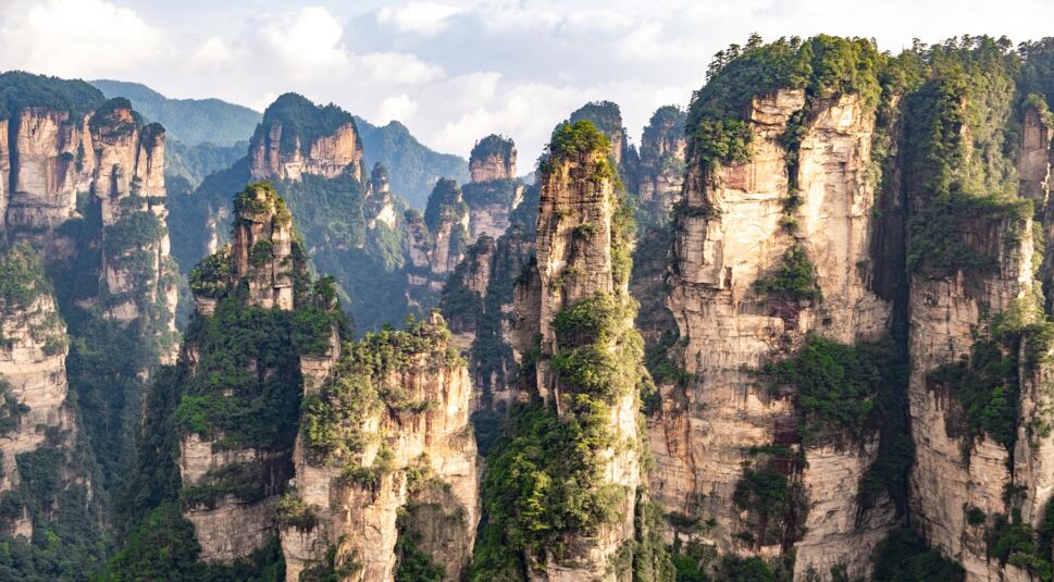 Auf dem Bild zu Avatar Fakten ist der Zhangjiajie National Forest Park in China zu sehen. Die beeindruckenden, säulenförmigen Sandsteinfelsen sind von dichtem, grünem Bewuchs umgeben und ragen majestätisch in den Himmel. Diese einzigartige Landschaft diente als Inspiration für die schwebenden Berge von Pandora im Film Avatar. Die Szene wirkt ruhig und atemberaubend zugleich.