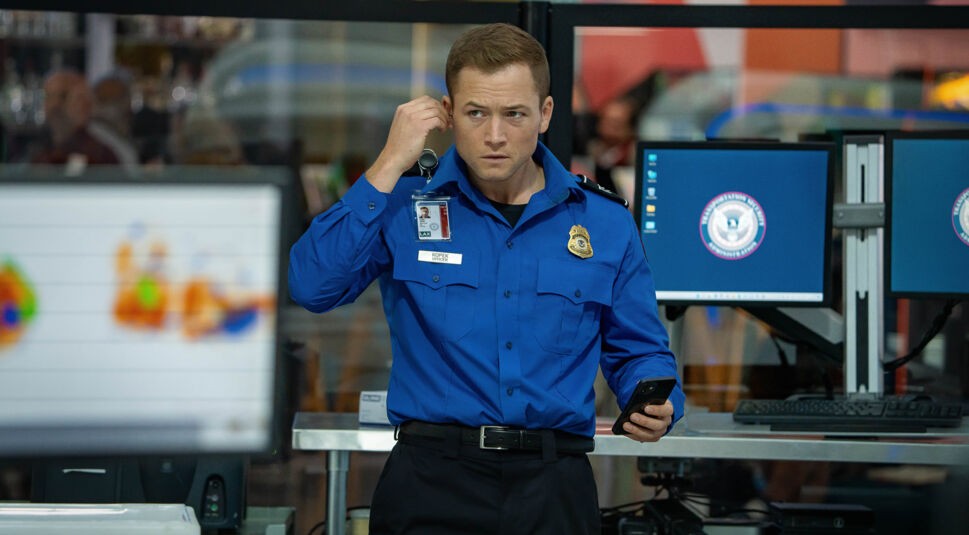 Auf dem Bild aus dem Film Carry-On ist Taron Egerton als Ethan Kopek in einer blauen TSA-Uniform zu sehen. Er trägt ein Namensschild und hält ein Kommunikationsgerät ans Ohr, während er in der anderen Hand ein Smartphone hält. Im Hintergrund sind Monitore mit offiziellen Logos und unscharfe Personen zu erkennen, die eine Arbeitsumgebung am Flughafen andeuten. Die Szene wirkt angespannt und geschäftig.