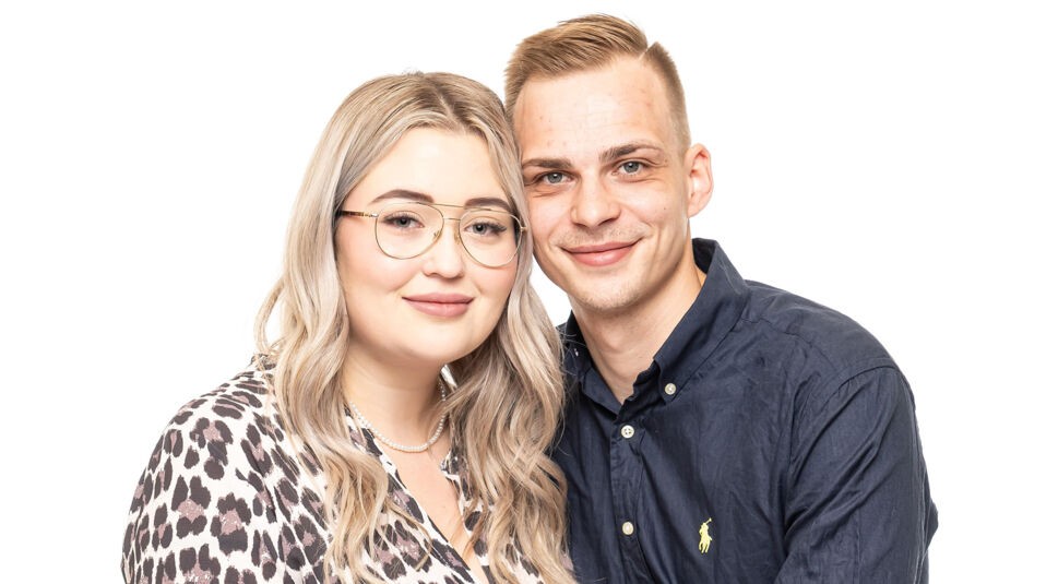 Auf dem Bild zu Das Sommerhaus der Normalos 2025 ist ein weiteres teilnehmendes Paar zu sehen. Die Frau hat lange, wellige blonde Haare, trägt eine Brille mit goldfarbenem Rahmen und ein gemustertes Oberteil. Der Mann hat kurze, hellbraune Haare und trägt ein dunkelblaues Hemd mit einem kleinen Markenlogo. Beide lächeln sanft und schauen direkt in die Kamera, während sie eng beieinanderstehen vor einem weißen Hintergrund.