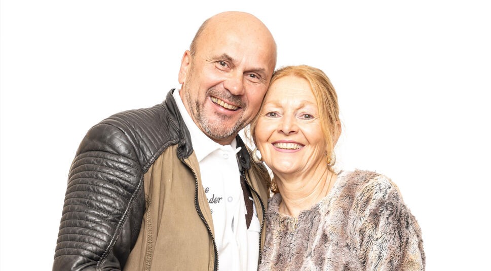 Auf dem Bild zu Das Sommerhaus der Normalos 2025 ist ein weiteres teilnehmendes Paar zu sehen. Der Mann hat eine Glatze, einen kurzen Bart und trägt eine zweifarbige Jacke mit schwarzen Lederärmeln über einem weißen Hemd. Die Frau hat rötlich-blonde Haare, die nach hinten gebunden sind, und trägt eine flauschige, gemusterte Jacke sowie goldene Ohrringe. Beide lächeln herzlich in die Kamera und stehen eng beieinander vor einem weißen Hintergrund.