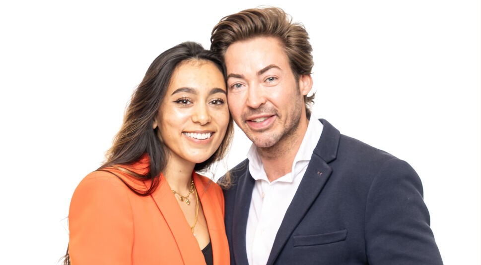 Auf dem Bild zu Das Sommerhaus der Normalos 2025 ist ein weiteres Paar der Show zu sehen. Die Frau hat lange, dunkle Haare und trägt einen auffälligen orangefarbenen Blazer mit goldenen Schmuckstücken. Der Mann hat volles, nach hinten gestyltes braunes Haar und trägt ein dunkles Sakko mit einem weißen Hemd. Beide lächeln in die Kamera und stehen eng beieinander vor einem weißen Hintergrund.