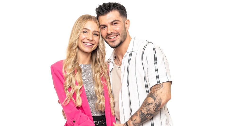 Auf dem Bild zu Das Sommerhaus der Normalos 2025 ist ein weiteres teilnehmendes Paar zu sehen. Die Frau hat lange, wellige blonde Haare und trägt einen pinken Blazer über einem silbernen, glitzernden Top. Der Mann hat dunkle, nach hinten gestylte Haare, einen kurzen Bart und trägt ein weißes Hemd mit schwarzen Streifen, das seine tätowierte Armmuskulatur betont. Beide lächeln in die Kamera und stehen eng beieinander vor einem weißen Hintergrund.