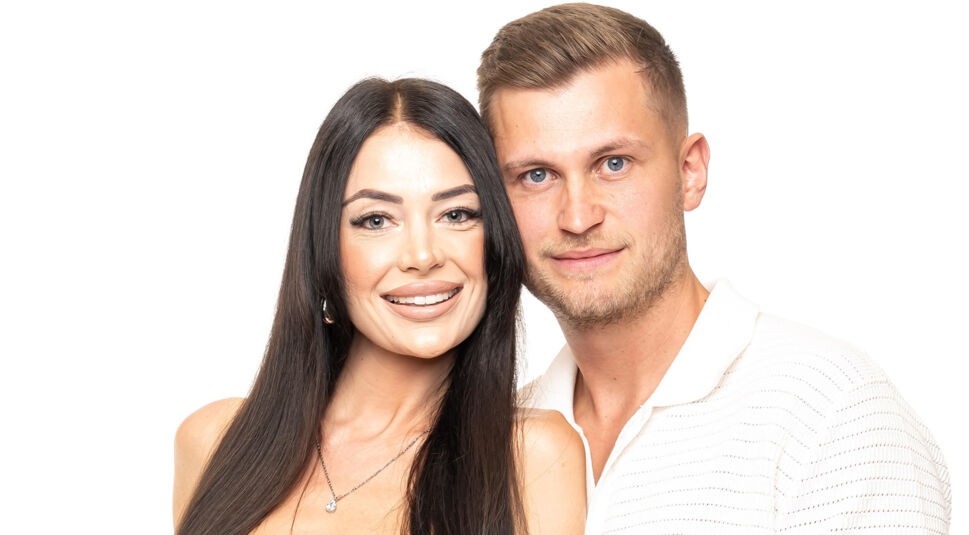 Auf dem Bild zu Das Sommerhaus der Normalos 2025 ist ein weiteres teilnehmendes Paar zu sehen. Die Frau hat lange, glatte schwarze Haare, trägt auffälliges Make-up mit betonten Lippen und Augen sowie eine silberne Halskette. Der Mann hat kurze, hellbraune Haare, blaue Augen und trägt ein weißes, leicht strukturiertes Poloshirt. Beide stehen eng beieinander, lächeln in die Kamera und posieren vor einem weißen Hintergrund.