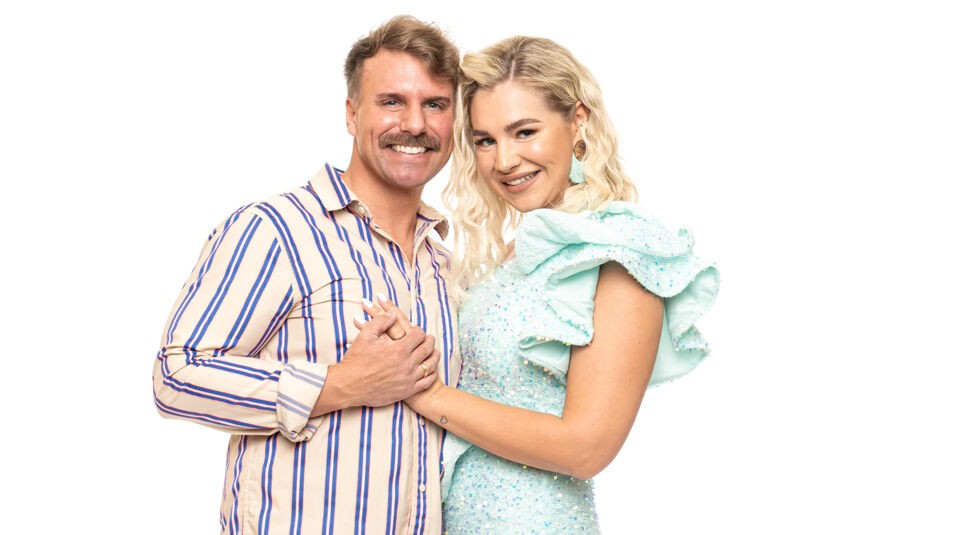 Auf dem Bild zu Das Sommerhaus der Normalos ist ein weiteres teilnehmendes Paar zu sehen. Der Mann hat kurze, wellige blonde Haare, einen markanten Schnurrbart und trägt ein beigefarbenes Hemd mit blauen und roten Streifen. Die Frau hat mittellange, wellige blonde Haare und trägt ein auffälliges, glitzerndes mintgrünes Kleid mit großen Rüschen. Beide halten sich an den Händen, lächeln in die Kamera und stehen eng beieinander vor einem weißen Hintergrund.