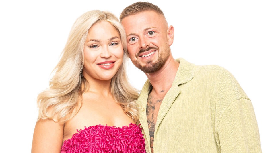 Auf dem Bild zu Das Sommerhaus der Normalos 2025 ist ein weiteres Paar der Show zu sehen. Die Frau hat lange, wellige blonde Haare und trägt ein auffälliges pinkes Kleid mit strukturierter Oberfläche. Der Mann hat kurze, braune Haare mit einem gestutzten Bart und trägt ein leicht geöffnetes, hellgrünes Hemd, das einige seiner Tattoos sichtbar macht. Beide lächeln in die Kamera und stehen eng beieinander vor einem weißen Hintergrund.