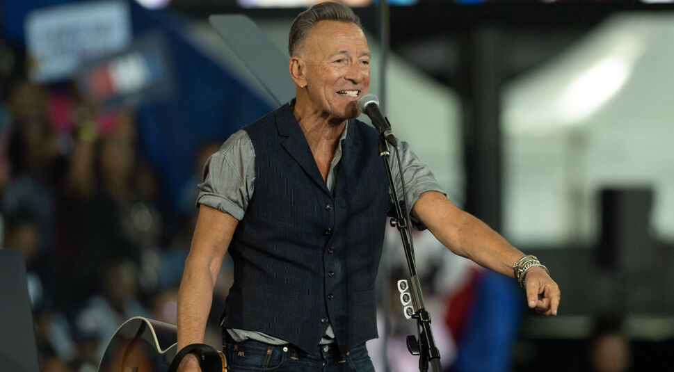 Auf dem Bild in dem Artikel über den Deliver Me From Nowhere-Film ist Bruce Springsteen zu sehen. Er steht auf einer Bühne und trägt ein graues Hemd mit hochgekrempelten Ärmeln sowie eine dunkle Weste. Vor ihm steht ein Mikrofon, und er hält eine Gitarre. Im Hintergrund sind unscharf Zuschauer:innen zu erkennen, die eine lebendige Konzertatmosphäre vermitteln.