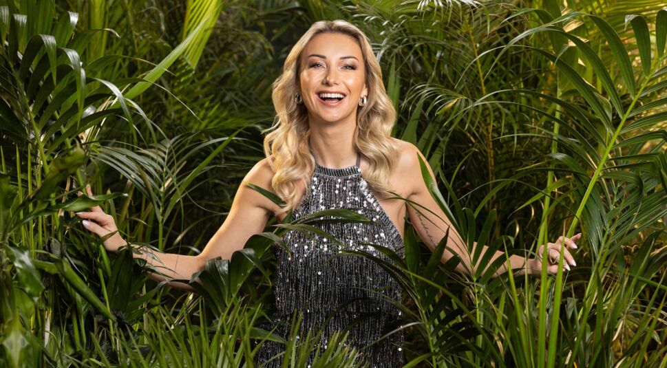 Auf dem Promobild zum Dschungelcamp 2025 ist Kandidatin Anna-Carina Woitschack zu sehen. Sie trägt ein glitzerndes silbernes Kleid mit Pailletten und steht lächelnd inmitten von üppigem tropischem Blattwerk.
