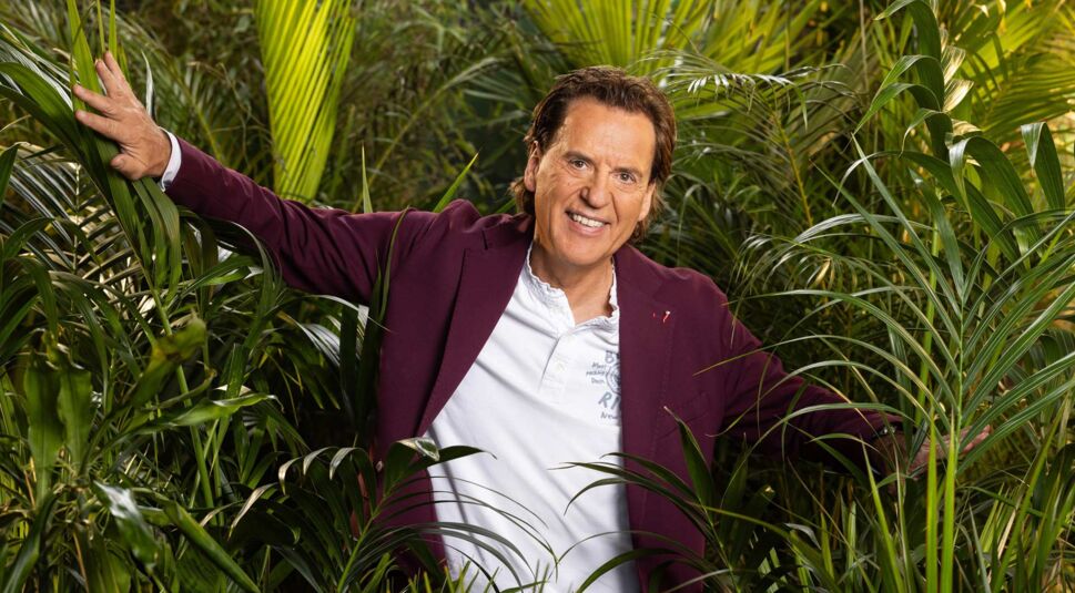 Auf dem Promobild zum Dschungelcamp 2025 ist Kandidat Jörg Dahlmann zu sehen. Er trägt ein weinrotes Sakko über einem weißen Poloshirt und steht inmitten von dichtem tropischem Blattwerk.