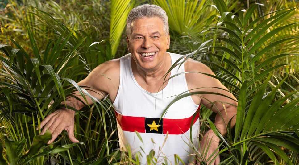 Auf dem Promobild zum Dschungelcamp 2025 ist Kandidat Jürgen Hingsen zu sehen. Er trägt ein weißes Tanktop mit einem roten Streifen und einem gelben Stern in der Mitte und steht inmitten von üppigem tropischem Blattwerk.