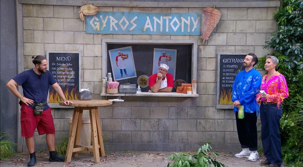 Auf dem Bild aus dem Dschungelcamp 2025 ist Maurice in einer Dschungelprüfung zu sehen. Er trägt ein dunkelblaues Shirt, rote Shorts und eine Bauchtasche, während er entspannt an einem Holztisch lehnt. Vor ihm steht ein Imbissstand mit der Aufschrift „Gyros Antony“, an dem ein Mann in Kochkleidung gelangweilt aus dem Fenster schaut. Rechts daneben stehen die Moderator:innen, gekleidet in auffällige bunte Outfits, und beobachten die Szene mit einem Lächeln.