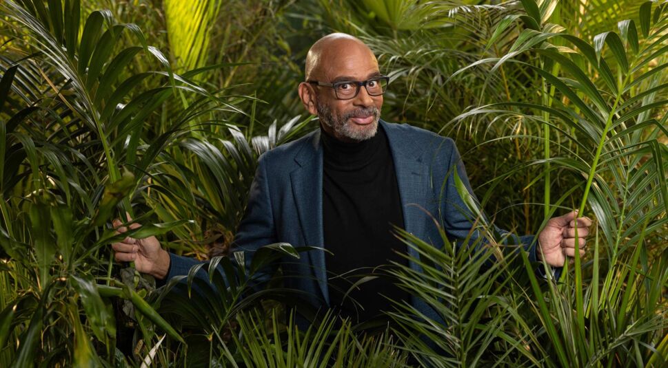 Auf dem Promobild zum Dschungelcamp 2025 ist Kandidat Pierre Sanoussi-Bliss zu sehen. Er trägt ein elegantes blaues Sakko über einem schwarzen Rollkragenpullover und steht inmitten von dichtem tropischem Blattwerk.