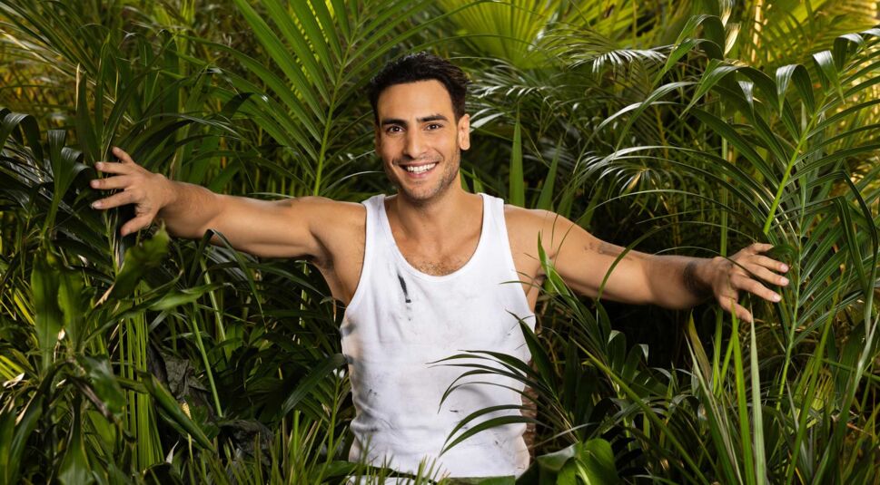 Auf dem Promobild zum Dschungelcamp 2025 ist Timur Ülker abgebildet. Er trägt ein weißes, leicht verschmutztes Tanktop und steht lächelnd inmitten von dichtem, tropischem Blattwerk.