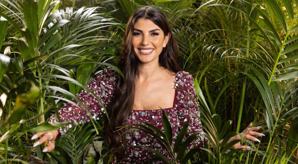 Auf dem Promobild zum Dschungelcamp 2025 ist Yeliz Koc zu sehen, umgeben von üppigem grünen Blattwerk. Sie trägt ein glitzerndes, mit Pailletten verziertes Kleid in Bordeauxrot und lächelt strahlend in die Kamera.