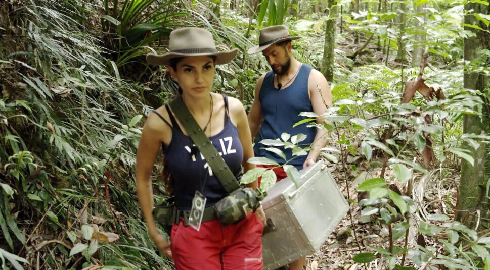 Auf dem Bild aus dem Dschungelcamp 2025 sind Yeliz und Maurice im Dschungel unterwegs. Yeliz läuft vorneweg, trägt einen Hut, ein dunkelblaues Top mit ihrem Namen und eine rote Hose. Maurice folgt ihr, ebenfalls mit Hut, in einem blauen Shirt und trägt eine große silberne Truhe. Die Umgebung ist grün und dicht bewachsen.