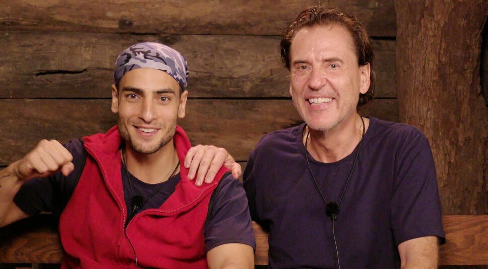 Auf dem Bild aus dem Dschungelcamp 2025 sind Jörg und Timur im Dschungeltelefon zu sehen. Timur trägt eine rote Weste über einem dunklen Shirt und ein gemustertes Bandana, während Jörg ein dunkelblaues Shirt trägt. Beide lächeln gut gelaunt in die Kamera, Timur ballt die Faust und Jörg legt die Hand auf seine Schulter. Die rustikale Holzwand im Hintergrund unterstreicht die typische Camp-Atmosphäre.