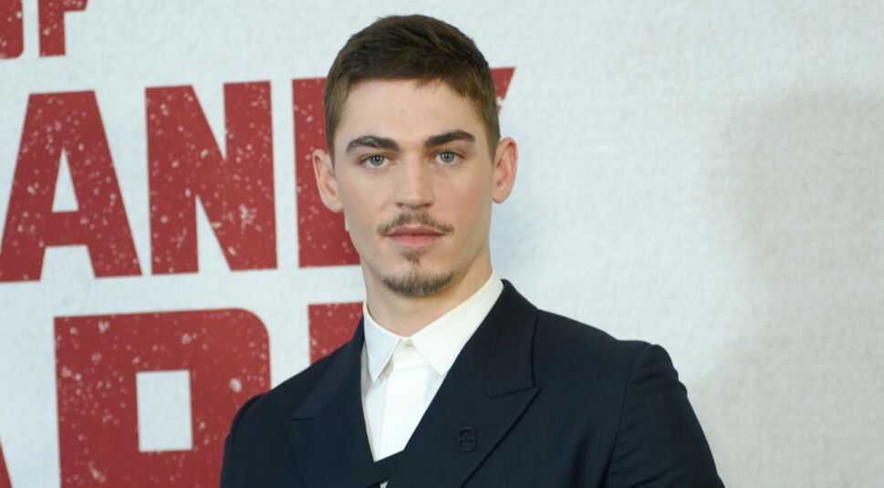 Hero Fiennes Tiffin ist auf diesem Bild vor einem weißen Hintergrund mit rotem Schriftzug zu sehen. Er trägt einen eleganten, dunklen Anzug mit weißem Hemd und schaut selbstbewusst in die Kamera. Sein Gesichtsausdruck ist ernst, aber aufmerksam. Dieses Foto steht im Kontext der "Fourth Wing Serie".