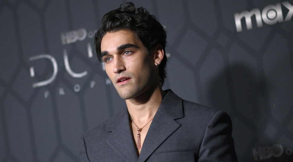 Josh Heuston ist auf diesem Bild vor einer dunklen Kulisse mit dem Schriftzug "HBO" und "Max" zu sehen. Er trägt einen eleganten, dunkelgrauen Anzug und eine auffällige Halskette mit einem goldenen Anhänger. Sein Blick ist nachdenklich und ausdrucksstark. Dieses Foto steht im Kontext der "Fourth Wing Serie".