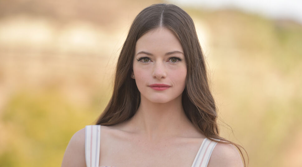 Mackenzie Foy ist auf diesem Bild in natürlichem Licht zu sehen, mit langen braunen Haaren, die offen über ihre Schultern fallen. Sie trägt ein helles, sommerliches Kleid mit dezenten Streifen. Der Hintergrund ist leicht unscharf und zeigt eine natürliche Umgebung. Dieses Foto steht im Zusammenhang mit der "Fourth Wing Serie".