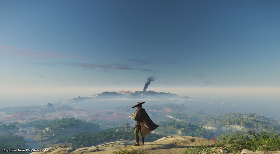 In diesem Bild aus Ghost of Tsushima steht ein Samurai mit traditionellem Hut und Umhang auf einem Hügel und blickt auf eine weitläufige Landschaft. Im Vordergrund erstrecken sich grüne Wälder und Dörfer, während im Hintergrund das Meer mit zahlreichen Segelbooten und eine Insel mit aufsteigendem Rauch zu sehen sind. Der klare blaue Himmel verleiht der Szene eine ruhige und friedliche Atmosphäre.