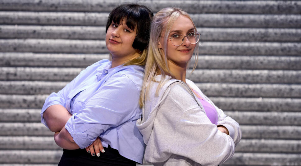 Auf dem Bild zu LEGO Masters 2025 sind die Kandidatinnen Feli und Lea zu sehen. Sie stehen Rücken an Rücken und verschränken selbstbewusst die Arme. Lea, links im hellblauen Hemd, hat dunkle Haare mit blondem Ombre-Effekt, während Feli, rechts, blonde Haare und eine Brille trägt. Sie trägt eine graue Kapuzenjacke über einem lila Oberteil. Der Hintergrund zeigt eine industrielle Metallstruktur.