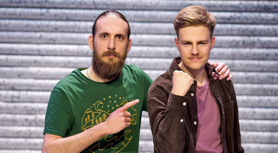 Auf dem Bild zu LEGO Masters 2025 sind die Kandidaten Filippos und Tobias zu sehen. Filippos, links im grünen T-Shirt, hat lange Haare und einen Vollbart. Er zeigt mit ernstem Blick auf Tobias, der rechts steht. Tobias trägt ein braunes Cordhemd über einem lila Shirt und ballt entschlossen die Faust. Die beiden wirken motiviert und stehen vor einem metallischen Hintergrund.