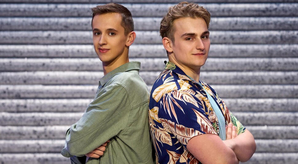 Auf dem Bild zu LEGO Masters 2025 sind die Kandidaten Leon und Marc zu sehen. Sie stehen Rücken an Rücken und haben die Arme verschränkt. Leon, links, trägt ein grünes Hemd und hat kurze braune Haare. Marc, rechts, trägt ein buntes Hawaii-Hemd mit Blattmuster und hat wellige blonde Haare. Beide blicken selbstbewusst in die Kamera. Der Hintergrund zeigt eine metallische Struktur.