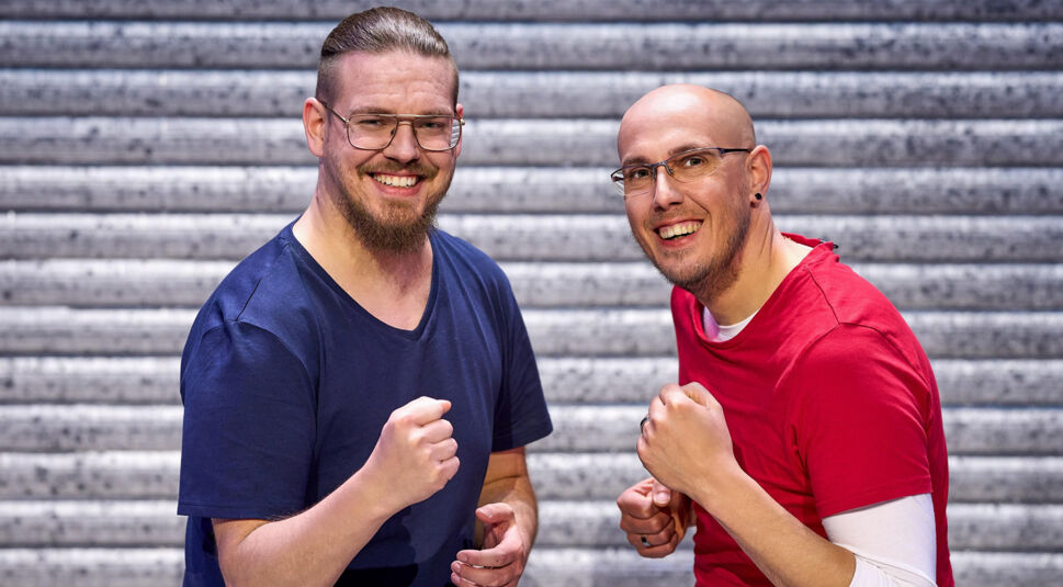 Auf dem Bild zu LEGO Masters 2025 sind die Kandidaten Mäkkes und Tobias zu sehen. Tobias, links, trägt ein dunkelblaues T-Shirt, hat einen Vollbart, nach hinten gebundene Haare und eine Brille. Mäkkes, rechts, trägt ein rotes Shirt über einem weißen Langarmshirt, ist glatzköpfig und trägt ebenfalls eine Brille. Beide lächeln und ballen spielerisch die Fäuste, während sie motiviert in die Kamera schauen. Der Hintergrund zeigt eine metallische Struktur.