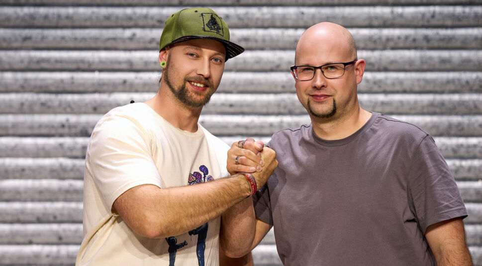 Auf dem Bild zu LEGO Masters 2025 sind die Kandidaten Sören und Sven zu sehen. Sören, links, trägt ein beiges T-Shirt mit einem bunten Print und eine grüne Cap. Er hat kurze Gesichtsbehaarung und grüne Tunnel-Ohrringe. Sven, rechts, trägt ein graues T-Shirt, ist glatzköpfig und hat eine Brille. Die beiden reichen sich die Hand in einem festen Griff und blicken entschlossen in die Kamera. Der Hintergrund zeigt eine metallische Struktur.