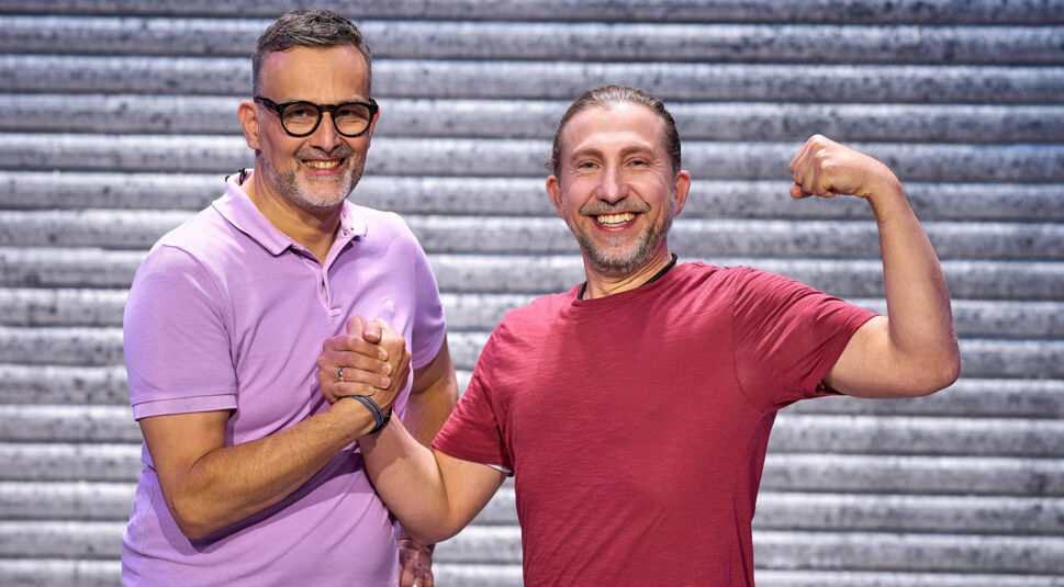 Auf dem Bild zu LEGO Masters 2025 sind die Kandidaten Thomas und Uwe zu sehen. Uwe, links, trägt ein lilafarbenes Poloshirt und eine Brille. Er hat kurze dunkle Haare mit grauen Ansätzen und einen gepflegten Bart. Thomas, rechts, trägt ein rotes T-Shirt, hat nach hinten gebundene Haare und ein breites Lächeln. Er hebt stolz seinen Arm und zeigt seinen Bizeps. Die beiden geben sich einen festen Händedruck und strahlen vor Freude. Der Hintergrund zeigt eine metallische Struktur.