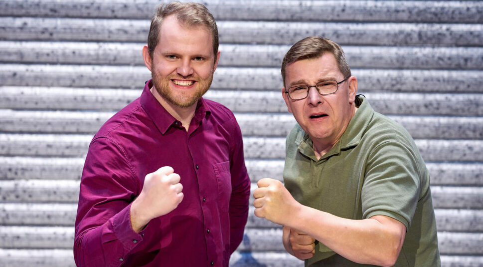 Auf dem Bild zu LEGO Masters 2025 sind die Kandidaten Tobias und Martin zu sehen. Martin, links, trägt ein bordeauxfarbenes Hemd, hat kurze blonde Haare und ein breites Lächeln. Tobias, rechts, trägt ein grünes Polohemd, eine Brille und macht ein humorvolles, angestrengtes Gesicht. Beide haben die Fäuste erhoben und wirken entschlossen. Der Hintergrund zeigt eine metallische Struktur.