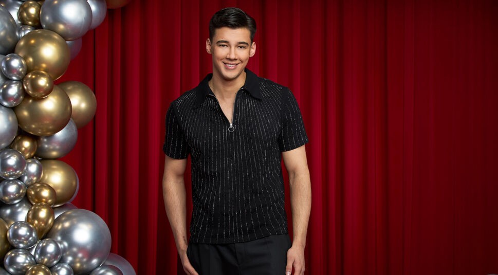 Auf dem Bild zu Let's Dance 2025 ist Diego Poth vor einem roten Vorhang zu sehen. Er trägt ein schwarzes, kurzärmliges Hemd mit glitzernden Streifen und einem Reißverschluss am Kragen. Seine dunklen Haare sind elegant gestylt, und er lächelt sympathisch in die Kamera. Links im Bild sind goldene und silberne Ballons dekorativ angeordnet, was eine festliche Atmosphäre schafft.