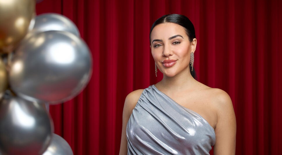 Auf dem Bild zu Let's Dance 2025 ist Leyla Lahouar vor einem roten Vorhang zu sehen. Sie trägt ein elegantes, silbernes One-Shoulder-Kleid mit glänzendem Stoff, das sanft fällt. Ihre dunklen Haare sind streng nach hinten gestylt, und sie trägt auffällige, funkelnde Ohrringe. Mit einem dezenten Lächeln blickt sie selbstbewusst in die Kamera. Links im Bild sind goldene und silberne Ballons dekorativ angeordnet.