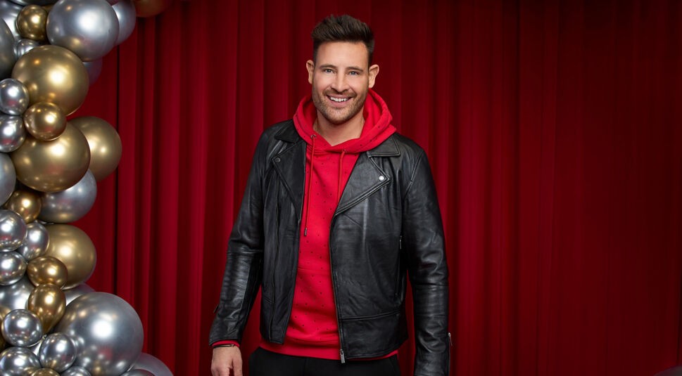 Auf dem Bild zu Let's Dance 2025 ist Marc Eggers vor einem roten Vorhang zu sehen. Er trägt einen roten Hoodie mit kleinen funkelnden Details unter einer schwarzen Lederjacke, was seinem Look eine lässige Note verleiht. Mit einem offenen Lächeln blickt er direkt in die Kamera. Links im Bild sind goldene und silberne Ballons dekorativ angeordnet, die eine festliche Atmosphäre schaffen.