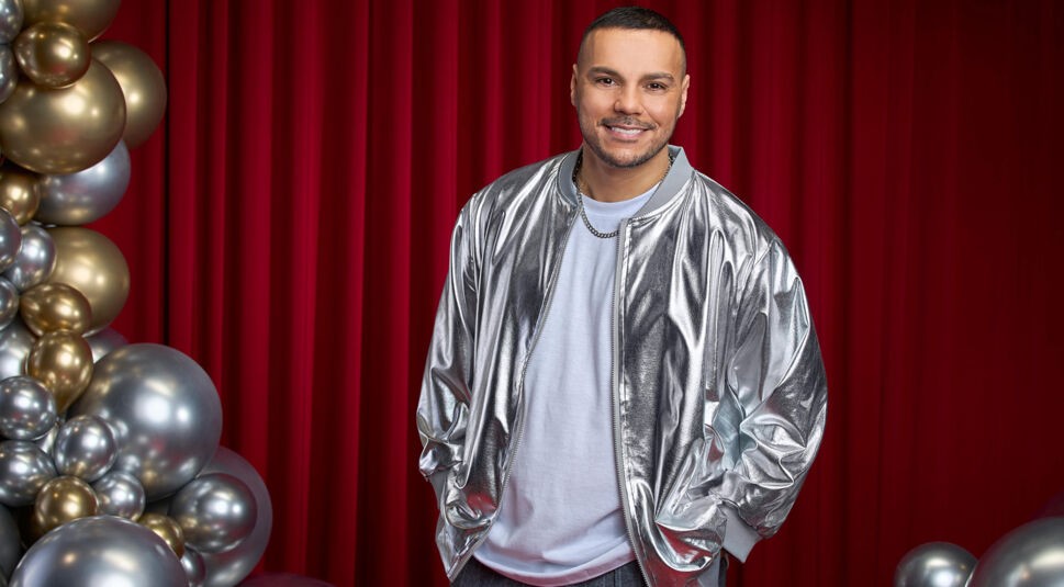 Auf dem Bild zu Let's Dance 2025 ist Osan Yaran vor einem roten Vorhang zu sehen. Er trägt eine auffällige silberne Bomberjacke über einem weißen Shirt und kombiniert den Look mit einer dezenten Silberkette. Mit entspannt in die Taschen gesteckten Händen und einem freundlichen Lächeln blickt er direkt in die Kamera. Links im Bild sind goldene und silberne Ballons dekorativ angeordnet, die eine festliche Atmosphäre schaffen.