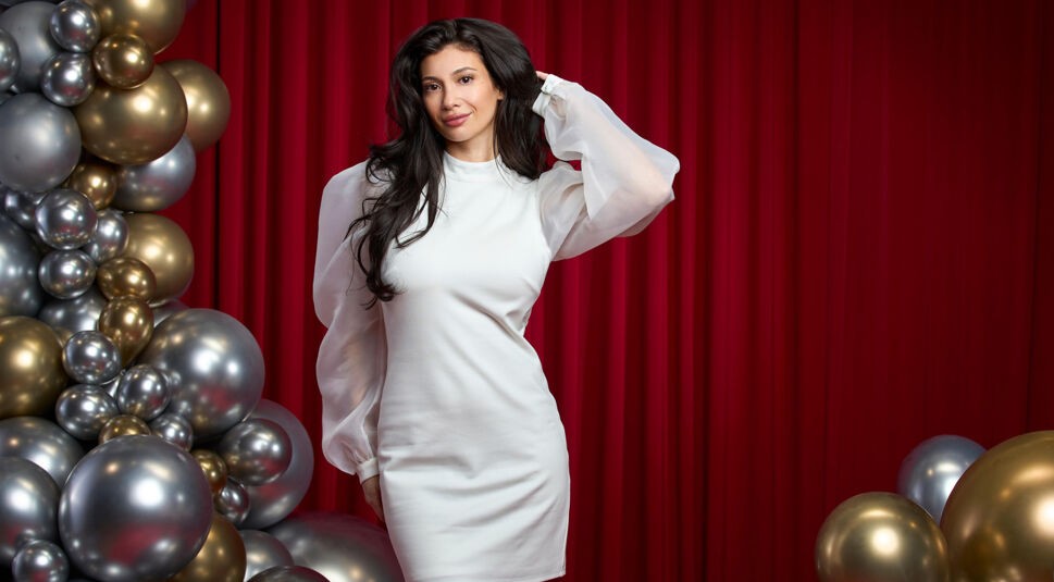 Auf dem Bild zu Let's Dance 2025 ist Paola Maria vor einem roten Vorhang zu sehen. Sie trägt ein elegantes, weißes Minikleid mit transparenten, leicht gebauschten Ärmeln. Ihre dunklen, langen Haare fallen in weichen Wellen über die Schultern, während sie mit einer Hand ihr Haar zurückstreicht. Mit einem sanften Lächeln blickt sie in die Kamera. Links im Bild sind goldene und silberne Ballons dekorativ angeordnet, die eine festliche Atmosphäre schaffen.