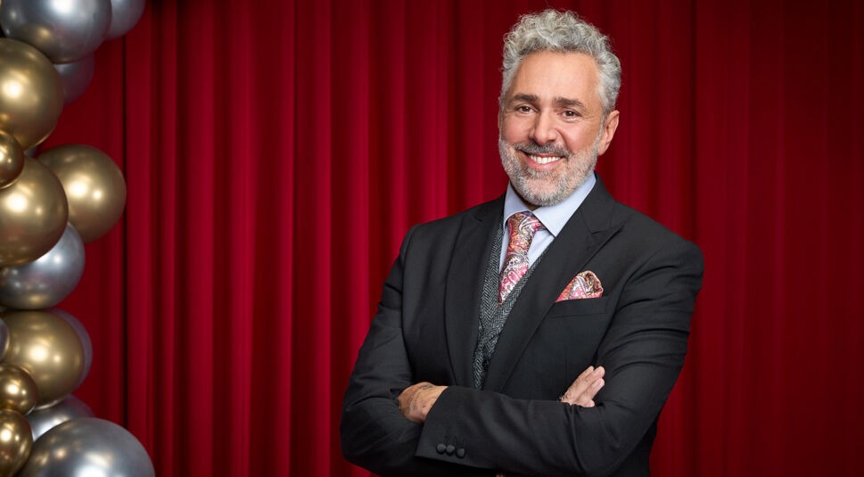 Auf dem Bild zu Let's Dance 2025 ist Roland Trettl vor einem roten Vorhang zu sehen. Er trägt einen eleganten schwarzen Anzug mit Weste, kombiniert mit einem hellblauen Hemd, einer gemusterten Krawatte und einem farblich passenden Einstecktuch. Seine grauen Locken und der Bart verleihen ihm einen markanten Look. Mit verschränkten Armen und einem freundlichen Lächeln blickt er in die Kamera. Links im Bild sind goldene und silberne Ballons dekorativ angeordnet.