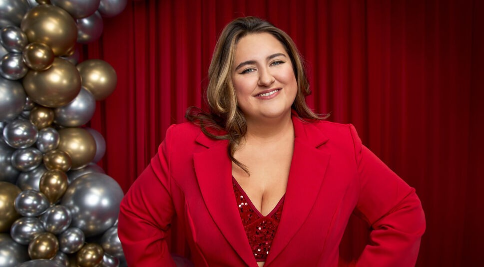 Auf dem Bild zu Let's Dance 2025 ist selfiesandra vor einem roten Vorhang zu sehen. Sie trägt einen auffälligen roten Blazer mit einem glitzernden roten Top darunter. Ihr welliges Haar fällt locker über die Schultern, und sie lächelt herzlich in die Kamera. Mit den Händen in der Taille wirkt ihre Pose selbstbewusst und strahlend. Links im Bild sind goldene und silberne Ballons dekorativ angeordnet, die eine festliche Atmosphäre schaffen.