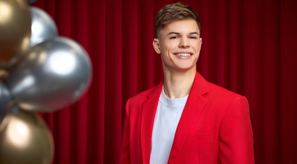 Auf dem Bild zu Let's Dance 2025 ist Taliso Engel vor einem roten Vorhang zu sehen. Er trägt einen leuchtend roten Blazer über einem schlichten weißen Shirt, was ihm einen modernen und stilvollen Look verleiht. Sein kurzes, blondes Haar ist locker gestylt, und er lächelt sympathisch in die Kamera. Links im Bild sind goldene und silberne Ballons dekorativ angeordnet, die eine festliche Atmosphäre schaffen.