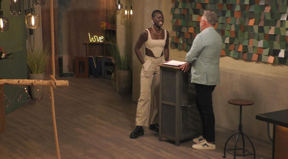 In diesem Bild aus „Promi First Dates“ ist Aminata Sanogo zu sehen, die entspannt und lächelnd vor einem Tresen steht. Sie trägt ein modernes, beigefarbenes Outfit mit Cut-Out-Details. Der Tresen wird von einem Mann in hellblauem Sakko und weißen Sneakern bedient. Im Hintergrund ist ein stilvoll dekorierter Raum mit einem leuchtenden „Love“-Schriftzug und einer bunten Wandgestaltung.