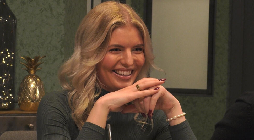 In diesem Bild ist Antonia Hemmer bei „Promi First Dates“ zu sehen. Sie lächelt herzlich und stützt ihr Kinn auf ihre verschränkten Hände. Sie trägt ein dunkelgraues Oberteil und eine Perlenarmkette. Im Hintergrund sind eine goldene Deko-Ananas und eine grüne Wand mit einem modernen Design zu erkennen, was eine elegante Atmosphäre schafft.