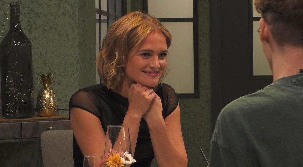 In diesem Bild aus „Promi First Dates“ ist Chiara Moon Horst zu sehen, wie sie mit einem Lächeln und gestütztem Kinn ihrem Gegenüber zuhört. Sie trägt ein schwarzes, leicht durchsichtiges Oberteil. Im Hintergrund befinden sich eine dekorative goldene Ananas und eine dunkelgrüne Wand.