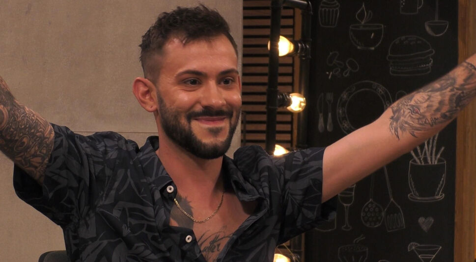 In diesem Bild aus „Promi First Dates“ ist Daniele Negroni zu sehen, wie er lächelnd die Arme ausbreitet. Er trägt ein dunkles Hemd mit tropischem Muster, das seinen tätowierten Armen Raum gibt. Auf seiner Brust ist ein weiteres Tattoo zu erkennen. Im Hintergrund sind stilisierte Zeichnungen von Geschirr und Küchenutensilien sowie warme Lichter zu sehen, die eine lockere Atmosphäre schaffen.