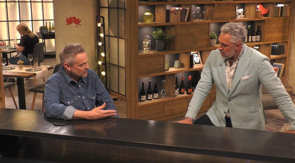 In diesem Bild aus „Promi First Dates“ ist Raphael Schneider zu sehen, wie er an der Bar in ein Gespräch mit dem Gastgeber im hellblauen Sakko vertieft ist. Raphael trägt ein lässiges Jeanshemd und gestikuliert leicht. Im Hintergrund sind stilvoll dekorierte Regale mit Büchern, Flaschen und Dekorationsobjekten zu sehen, die eine gemütliche Atmosphäre schaffen. Links sitzt eine weitere Teilnehmerin an einem Tisch.