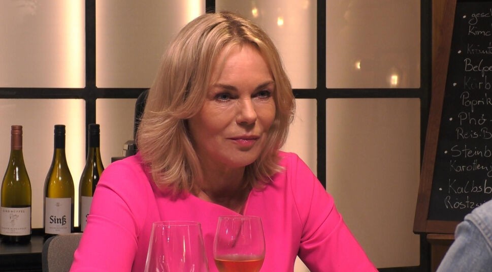 In diesem Bild aus „Promi First Dates“ ist Saskia Valencia zu sehen, wie sie aufmerksam ihrem Gegenüber zuhört. Sie trägt ein auffälliges pinkfarbenes Oberteil und ihr blondes Haar ist schulterlang gestylt. Auf dem Tisch vor ihr stehen zwei Gläser. Im Hintergrund sind Flaschen und eine Tafel mit handgeschriebenen Gerichten zu erkennen, was eine stilvolle und entspannte Atmosphäre schafft.